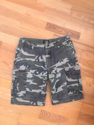 Cargoshorts i camo från Dickies w32 - Säljer ett par cargoshorts från Dickies i klassiskt camouflagemönster. Shortsen har flera praktiska fickor på sidorna och är tillverkade i ett slitstarkt bomullstyg. Perfekta för dig som gillar streetwear och utilitystil.
