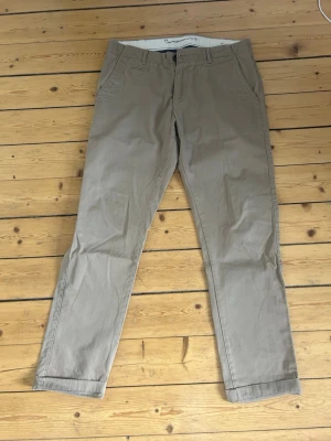 Beige chinos från Knowledge Cotton - Snygga beige chinos från Knowledge Cotton Apparel med klassisk rak passform. Byxorna har fickor fram och bak, bälteshällor och är tillverkade i mjuk bomull. Perfekta för en stilren och avslappnad look.
