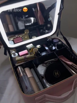 Rosa, svart sminkväska med spegel - Supersöt rosa sminkväska i fyrkantig form med guldfärgade dragkedjor och handtag. Väskan har inbyggd spegel med LED-belysning och flera smarta fack för att organisera smink och accessoarer. Materialet ser ut att vara konstläder.