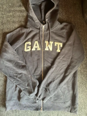 Mörkgrå zip-hoodie från GANT S/M - Snygg mörkgrå hoodie från GANT med dragkedja och stor huva. Klassisk logga i gult och vitt broderat på bröstet. Mjuk insida och ribbade muddar vid ärmar och nederkant. Perfekt för en avslappnad och sportig stil. Står XL i storlek men känns som en S/ M