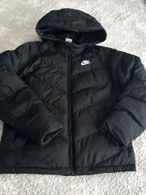 Svart pufferjacka från Nike 147-158 cm  - Svart pufferjacka från Nike med huva och dragkedja framtill. Jackan har ett vitt Nike-logga på bröstet och är vadderad för extra värme. Perfekt för kalla vinterdagar och har en klassisk, sportig look.