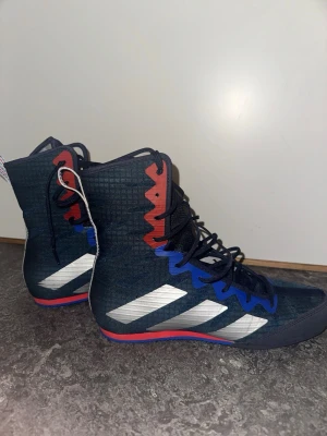 Adidas brottarskor blå/röd - Säljer ett par höga brottarskor från Adidas i mörkblått med silvriga ränder på sidorna. Skorna har svarta snören, blå och röda detaljer vid snörningen samt röd och blå sula. Perfekta för träning eller tävling inom brottning.