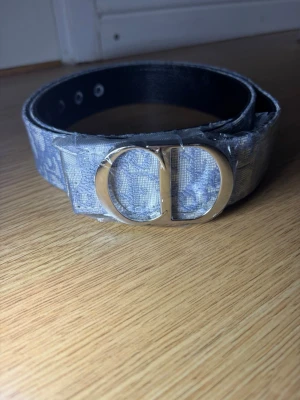 Grått bälte med silverfärgat spänne - Snyggt grått bälte med mönstrad textil och ett stort, silverfärgat spänne i form av bokstäverna CD. Bältet har flera hål för justering och insidan är svart. Perfekt accessoar för att lyfta din outfit.