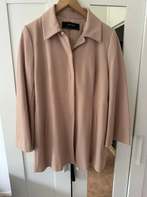 Beige rock från Zara Basic - Stilren beige rock från Zara Basic med klassisk krage och dolda knappar framtill. Rak passform och två diskreta fickor på sidorna. Perfekt för dig som gillar minimalistisk och tidlös stil. Materialet känns mjukt och har en lätt struktur.