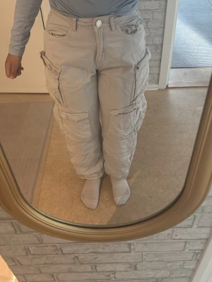 Beiga cargobyxor med fickor - Säljer ett par beiga cargobyxor med flera stora fickor på sidorna och avslappnad passform. Byxorna har knapp och dragkedja framtill samt detaljerade sömmar. Kommer självklart strykas innan den skickas