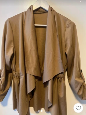 Beige trenchcoat med draperad front - Stilren beige trenchcoat med draperad öppen front och resår i midjan för en snygg siluett. Jackan har långa ärmar med knappdetalj för uppvik och är tillverkad i ett mjukt, lätt tyg som ger ett avslappnat intryck.