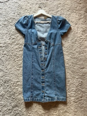 Blå jeansklänning med knappar från Levis  - Snygg blå jeansklänning med korta puffärmar och djup v-ringning. Klänningen har knappar framtill hela vägen ner och markerad midja för en feminin siluett. Perfekt till sommaren och enkel att styla med sneakers eller boots.