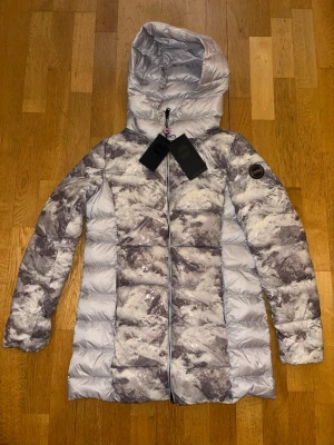 Colmar Originals dunjacka med huva – Ny med prislapp – Strl 42 - Snygg och exklusiv Colmar Originals dunjacka med huva i ett stilrent grått mönster. Lätt, varm och bekväm med hög kvalitet och modern passform.  Ny med prislapp.  * Märke: Colmar Originals * Storlek: 42 * Färg: Grå/mönstrad * Huva  