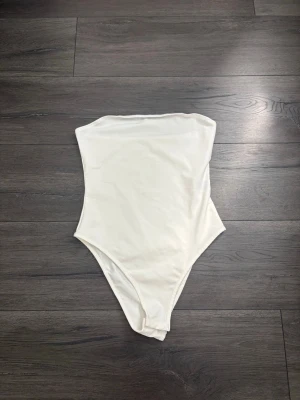 Vit axelbandslös body - Säljer en stilren vit body utan axelband. Bodyn har en enkel och tight passform med hög skärning vid benen. Tillverkad i ett stretchigt och mjukt material som sitter skönt mot kroppen. Perfekt att styla med jeans eller kjol.