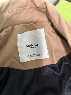 Beige trenchcoat från Mango - Stilren beige trenchcoat från Mango med dubbelknäppning och breda slag. Jackan har två framfickor och är tillverkad i ett mjukt, vävt material. Perfekt oversized passform för en avslappnad look.
