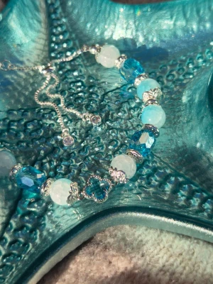 Tidal Whisper 🌊 Armband - Snyggt armband med blåskiftande glaspärlor i 8 mm och facetterade skimrande glaspärlor, dekorerat med glittrande silverfärgade detaljer och en liten fyrklöverformad dekoration i mitten. Armbandet har justerbar kedja med små stenar i ändarna. Perfekt accessoar för att lyfta din stil.