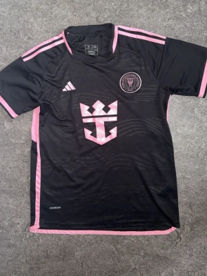 Svart Inter Miami matchtröja - Snygg svart matchtröja från Inter Miami med rosa detaljer och klubbmärke på bröstet. Tröjan har korta ärmar, rund hals och är tillverkad i ett lätt funktionsmaterial. Adidas-logga på framsidan och rosa ränder på axlarna ger en sportig look.