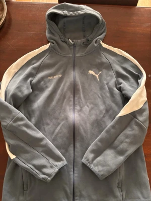 Blå hoodie Malmö FF Puma - Snygg blå hoodie från Puma med Malmö FF-tryck på bröstet och vita detaljer längs ärmarna. Dragkedja framtill, huva med dragsko och två fickor. Perfekt för dig som gillar sportig stil och vill representera ditt lag. Materialet känns mjukt och bekvämt.