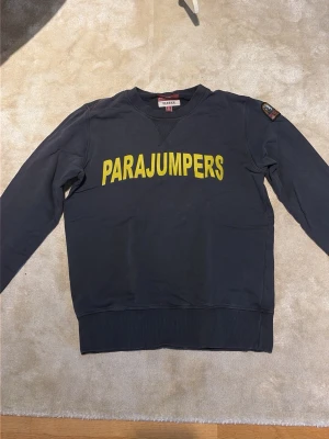Mörkblå sweatshirt från Parajumpers - Mörkblå sweatshirt från Parajumpers med gul logotyptext framtill. Tröjan har rund halsringning, ribbade muddar vid ärmslut och nederkant samt en patch på ena ärmen. Tillverkad i mjuk fleece för extra komfort.