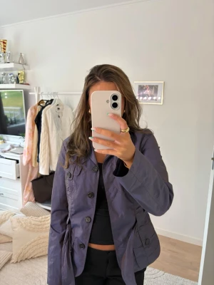 Lila/blå trenchcoat  - Snygg lila/blå trenchcoat med knytskärp i midjan och stora knappar framtill. Köpt ifrån sellpy men kommer ifrån united colors of benetton.
