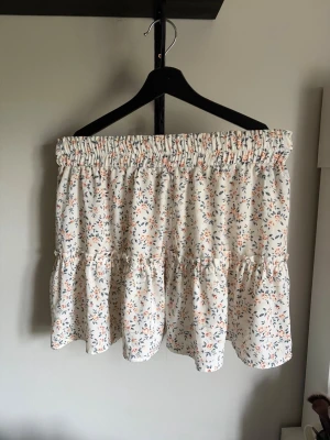 Vit blommig volangkjol - Söt kort kjol i vitt med små blommor i orange och blått. Kjolen har bred resår i midjan och volangdetaljer som ger en luftig och somrig känsla. Perfekt för dig som gillar en feminin och avslappnad stil.