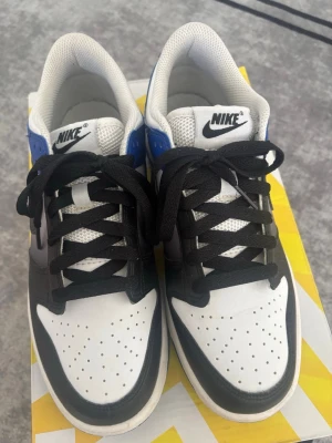 Nike Dunk Low svart/blå/vit - Nike Dunk Low sneakers i svart, blått och vitt med klassisk Swoosh på sidan. Ovandelen är i läder med perforerad tåbox för ventilation, svart snörning och vit mesh på plösen. Yttersulan är blå och ger en snygg kontrast till resten av skon.