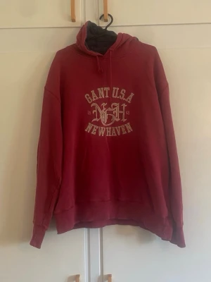 Röd hoodie GANT USA New Haven - Röd hoodie från GANT med tryck 'GANT U.S.A New Haven' på bröstet och dragsko i huvan. Klassisk passform med ribbade muddar vid ärmar och nederkant. Perfekt för en avslappnad och sportig stil.