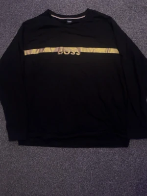 BOSS långärmad tröja i svart - BOSS långärmad tröja i svart med guldfärgad logotyp framtill. Stilren och bekväm modell som passar både till vardag och en mer uppklädd look.