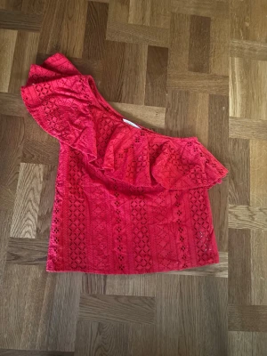 Röd spetsig oneshoulder topp - Snygg röd oneshoulder topp med volang och spetsdetaljer. Toppen har ett unikt mönster i spets och är axelbandslös på ena sidan. Perfekt för dig som vill sticka ut med en färgstark och trendig look.