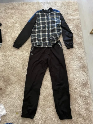Rutigt träningsset från Lacoste - Snyggt träningsset från Lacoste med svart-rutigt mönster i blått, vitt och grönt på jackan samt svarta ärmar och byxor. Setet har dragkedja framtill och klassisk Lacoste-logga på både jacka och byxa. Perfekt för sport eller chill.