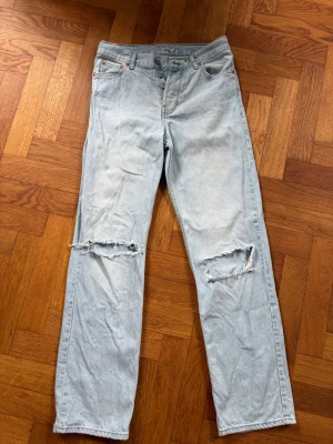 Ljusblå raka jeans med hål - Säljer ett par ljusblå raka jeans från Junkard Nevermind i storlek 28. Jeansen har slitna hål över båda knäna och klassisk knappgylf. Perfekta för en avslappnad och trendig look.
