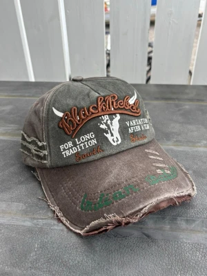 Black rebel distressed cap brun Grön vintage keps med brodyr - Säljer en brun och grön keps med slitna detaljer och broderad text 'BlackRebel' samt motiv av en tjurskalle. Kepsen har även broderade texter i vitt, orange och grönt samt dekorativa sömmar på sidorna. Justerbar baktill och tillverkad i bomull.