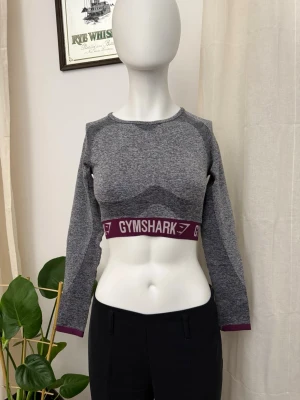 Grå croppad topp från Gymshark - Snygg grå croppad topp från Gymshark med lång ärm och bred resår under bysten i vinrött med vit logga. Tillverkad i stretchigt syntetmaterial, perfekt för träning. Sportig och modern look med rund halsringning.