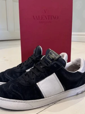 Valentino Garavani svarta sneakers - Snygga svarta sneakers från Valentino Garavani med vit panel på sidan och vita detaljer. Skorna har svart mocka och läder, klassisk låg modell och coola nitar på sulan. Perfekta för dig som vill ha en stilren och exklusiv look.