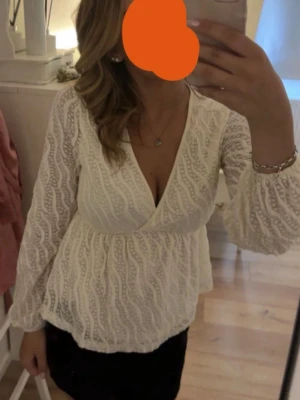 Vit omlottblus med spetsdetaljer - Snygg vit omlottblus med vågiga spetsdetaljer och djup v-ringning. Blusen har långa, lätt utsvängda ärmar och ett luftigt, romantiskt fall. Perfekt att styla med kjol eller jeans för en trendig look.