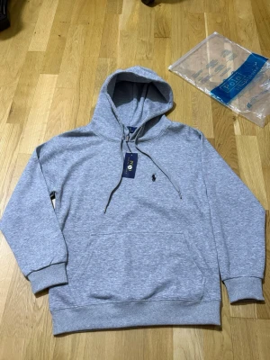 Grå hoodie från Polo Ralph Lauren - Snygg grå hoodie från Polo Ralph Lauren med klassisk huva och dragsnören. Broderad logga på bröstet och stor känguruficka framtill. Mjuk bomullsblandning och ribbade muddar vid ärmslut och nederkant. Perfekt för en chill och stilren look.