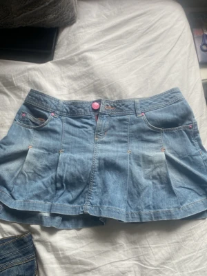 Ljusblå jeanskjol med rosa detaljer - Säljer en kort ljusblå jeanskjol med utsvängd passform och rosa detaljer på knappar och sömmar. Kjolen har fickor fram och bak samt en knappstängning framtill. Perfekt för dig som gillar en avslappnad och trendig stil.