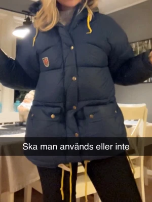 Mörkblå dunjacka från Fjällräven - Mörkblå dunjacka från Fjällräven med gula snören i huvan, stora fickor med knappar och klassisk logga på bröstet. Jackan har dragkedja och tryckknappar framtill samt justerbar nederkant. Perfekt för kalla vinterdagar. Storlek xxs MEN den är stor i storleken, den passar mig som är s vanligtvis 