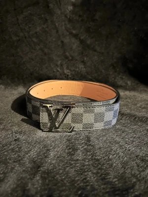 Rutigt L-bälte - Säljer ett stilrent bälte med klassiskt rutigt mönster i grått och svart samt en stor L-logga i metall som spänne. Bältet har en lyxig känsla och passar perfekt till jeans eller kostymbyxor.
