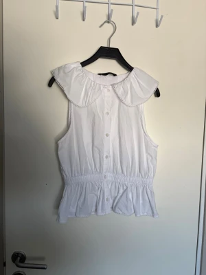 Vit ärmlös blus med volangkrage - Söt vit blus i bomull med bred volangkrage och knappar framtill. Modellen är ärmlös och har resår i midjan som ger en lätt rynkad effekt och volang nedtill. Perfekt för en fräsch och somrig look.