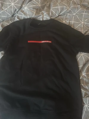 Svart sweatshirt från Prada - Snygg svart sweatshirt från Prada med röd logga och vit text på bröstet. Tröjan har rund halsringning och är tillverkad i mjukt material som känns skönt mot huden. Perfekt för en stilren och trendig look.