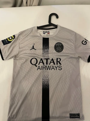 PSG Mbappé matchtröja grå - Snygg grå PSG matchtröja med svart tryck, Qatar Airways sponsor och Mbappé nummer 7 på ryggen. Tröjan har korta ärmar, svart rand i mitten och Paris Saint-Germain-logga på bröstet. Tillverkad i lätt och ventilerande material, perfekt för fotbollsträning eller match.