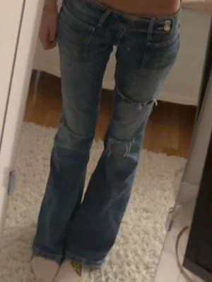 Low waist Blå bootcut jeans med slitningar - Säljer ett par blå lågmidjade  bootcut jeans med låg midja och slitningar på benen. Jeansen har klassisk femficksdesign och stängs med knapp och dragkedja. Perfekta för en avslappnad och trendig look. 