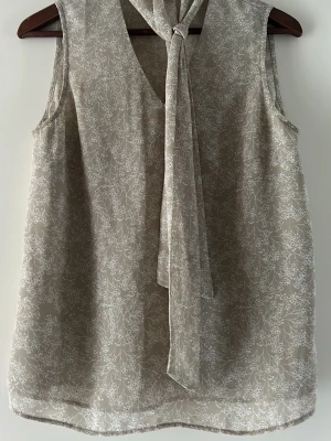 Beige blommig ärmlös blus H&M - Säljer en beige ärmlös blus från H&M med vitt blommönster och knytband vid halsen. Blusen har en lätt och luftig känsla, perfekt för varmare dagar. Passar dig som gillar en feminin och stilren look.