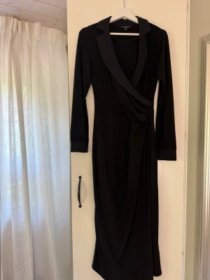 Svart omlottklänning med krage - Stilren svart långärmad omlottklänning med klassisk krage och knytning i midjan. Klänningen har en elegant siluett och är vadlång. Perfekt för dig som gillar en tidlös och chic look. Jätteskön klänning som sitter väldigt mjukt på kroppen☺️