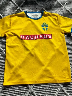 Svensk gul fotbollströja Bauhaus sponsorlogga - Gul fotbollströja med blå detaljer och Sveriges landslagsemblem på bröstet. Bauhaus-logga tryckt både fram och bak samt stort nummer 19 på ryggen. Perfekt för match eller supporterhäng. Rund halsringning och korta ärmar. Lite slitet på framsidan, bild 5.