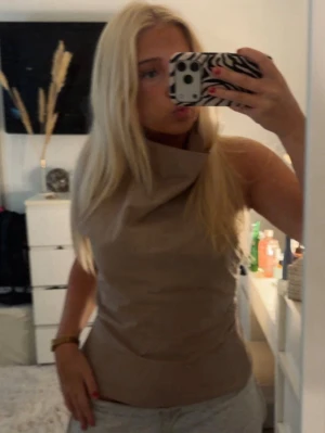 Beige ärmlös topp med hög krage - Säljer en beige ärmlös topp med hög, draperad krage och asymmetrisk halsringning. Toppen har en slim passform och är tillverkad i ett slätt material som ger en stilren look. Perfekt för dig som vill ha något trendigt och unikt.