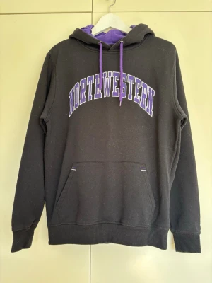 Svart hoodie med lila NORTHWESTERN-tryck, storlek M herr - Säljer en svart hoodie med lila huva och snören samt stort tryck 'NORTHWESTERN' i lila och vitt på bröstet. Hoodien har känguruficka framtill och ribbade muddar vid ärmar och nederkant. Storlek M herr. 70% bomull/ 30% polyester 