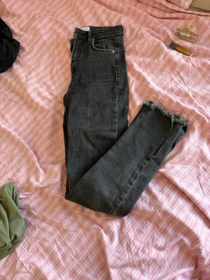 Grå jeans med rå kant från ZARA - Säljer ett par grå jeans från ZARA i storlek 34. Jeansen har hög midja, klassisk femficksdesign och rå, fransig kant vid bensluten för en trendig look. Perfekta till sneakers eller boots och passar till många olika stilar.