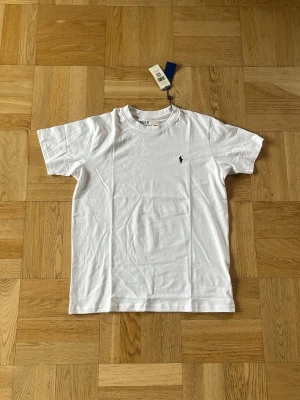 Vit t-shirt från Polo Ralph Lauren - Klassisk vit t-shirt från Polo Ralph Lauren med den ikoniska broderade loggan på bröstet. T-shirten har rund halsringning och korta ärmar. Tillverkad i mjuk bomull för en skön känsla och enkel stil.