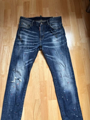 dsquared2 - Säljer ett par mörkblå jeans med slitningar, hål på ena knät och coola färgstänk i vitt och rött. Jeansen har en smal passform och klassisk femficksdesign. Materialet är denim med en tvättad look och detaljer som ger en trendig och avslappnad vibe.
