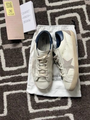 Vita sneakers med blå detalj - Snygga vita sneakers från Golden Goose med grå stjärna på sidan och blå hälkappa. Skorna har vita snören, beige sula och är tillverkade i läder med mockadetaljer. Perfekta för en trendig och avslappnad stil.
