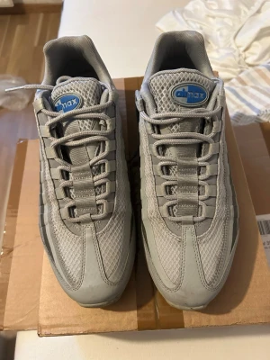 Nike Air Max 95 grå sneakers - Nike Air Max 95 sneakers i grått med blå detaljer. Ovandelen är i mesh och syntetmaterial med klassiska vågade paneler och synlig Air Max-logga på plösen. Skorna har en chunky sula och snörning framtill för en sportig look.
