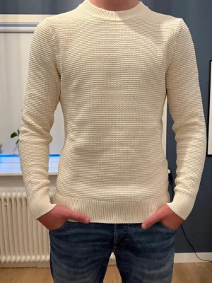 Vit finstickad tröja - Snygg vit finstickad tröja med rund hals och långa ärmar. Tröjan har en snygg passform med ribbade muddar vid ärmslut och nederkant. Perfekt att matcha med jeans för en clean och stilren look.