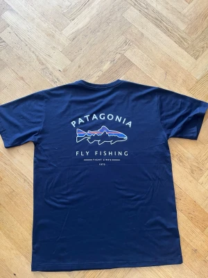Mörkblå Patagonia t-shirt med fisktryck - Snygg mörkblå t-shirt från Patagonia med tryck av en fisk och texten 'Fly Fishing' på ryggen  passar M -L